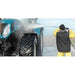 A person in a black vest, yellow pants, and black beanie uses a pressure washer on the side of a teal agricultural tractor, with water spraying in the air.