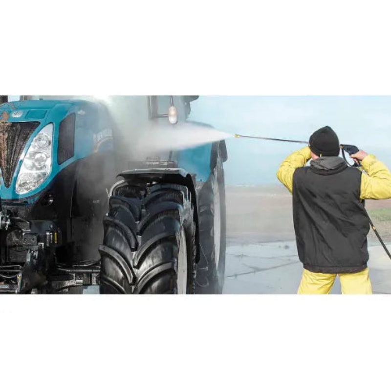 A person in a black vest, yellow pants, and black beanie uses a pressure washer on the side of a teal agricultural tractor, with water spraying in the air.