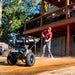 A man in a red plaid shirt and jeans uses a high-pressure water hose to wash a concrete driveway, with a black and silver pressure washer cart parked in the foreground on a sunny residential property.